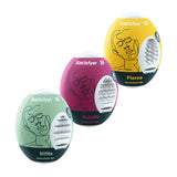 The Satisfyer Masturbator Eggs - Mixed 3 Pack #1 showcases three egg-shaped stroker sleeves: Riffle (green), Bubble (pink), and Fierce (yellow). Each features an abstract face outline, revealing the cyber-skin textured interior. The word Masturbator is below each name, with the Satisfyer logo on top.