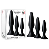 A set of three black silicone butt plugs, part of the Adam & Eve Silicone Booty Boot Camp Training Kit, offering graduated sizes for anal exploration, sits next to its branded packaging for varying levels from beginner to expert.