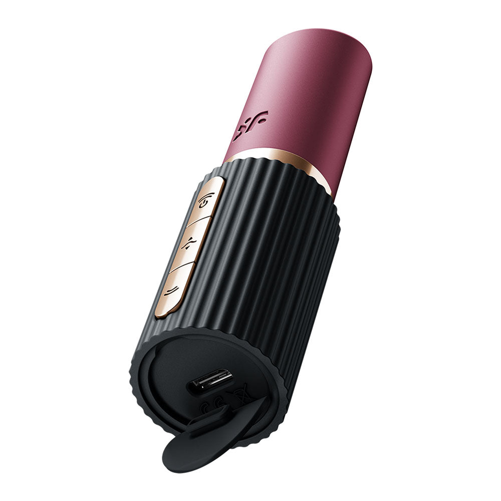 The Satisfyer Pro 2 Kiss is a cylindrical air pulse stimulator with a ribbed black base and sleek maroon top. It has gold buttons featuring plus, minus, and play symbols, a USB charging port covered with rubber on the bottom, and the brand logo on the maroon section.