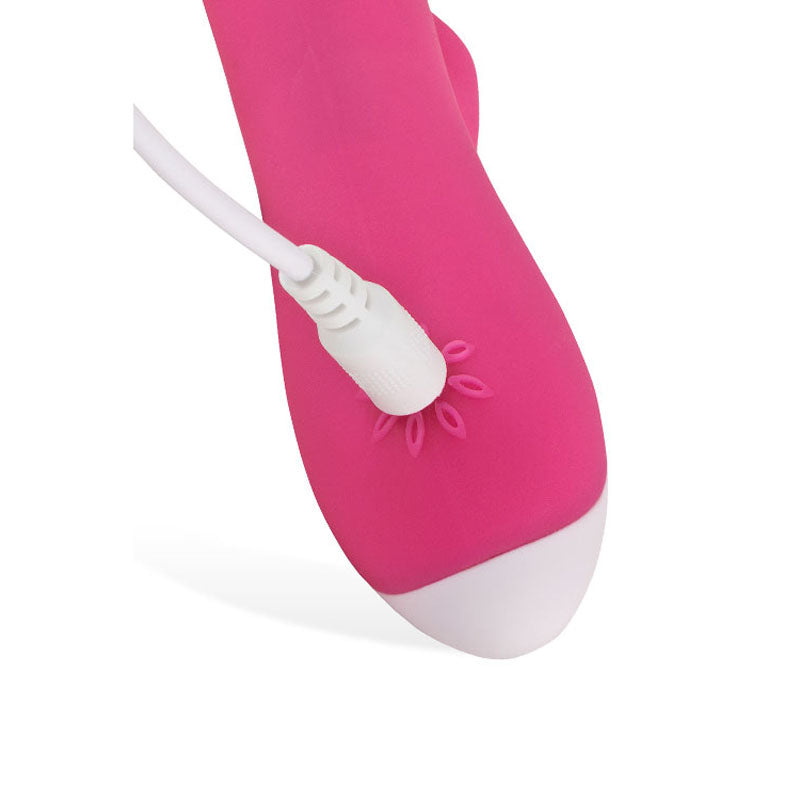 A close-up showcases the Adam & Eve The Warming Rabbit G USB Rechargeable Vibrator in pink, featuring a detachable white power cord. The charging port is surrounded by petal-like accents, and the device tapers to a white tip, designed for dual stimulation against a plain white background.