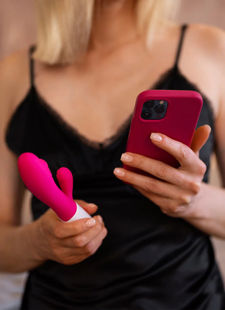 A person in a black lace-trimmed camisole holds a bright pink mobile phone in one hand and a pink silicone vibrating device in the other. The background is soft-focused, highlighting the persons nail polish and the vibrancy of the objects.