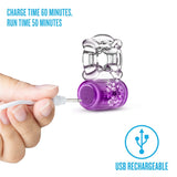 A purple Play With Me Pleaser Rechargeable C-Ring is being charged by hand. It boasts powerful vibrations, a 60-minute charge time, and 50-minute run time. USB rechargeable, with a USB symbol nearby.