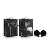 Two black boxes with NOIR branding feature elegant geometric designs and a hand graphic. Beside them are circular, opulent black fur-covered nipple clamps with metallic accents. These nickel-free Noir Pom Adjustable Nipple Clamps enhance the sleek, luxurious vibe.