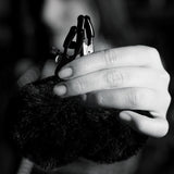 Close-up of a hand holding black fuzzy handcuffs, their texture highlighted by monochromatic lighting. Neatly manicured nails add elegance to this sensation play accessory, similar to Noir Pom Adjustable Nipple Clamps - Black Furry Nipple Clamps. A blurred background spotlights the cuff details.