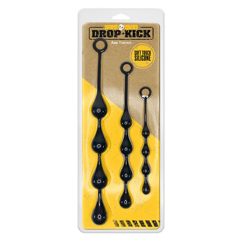 The packaging for the Boneyard Drop-Kick Ass Trainers showcases a set of three black silicone anal beads with round handles through its clear cover, against a brown background highlighted with Soft Touch Silicone and yellow accents for enhanced pleasure.