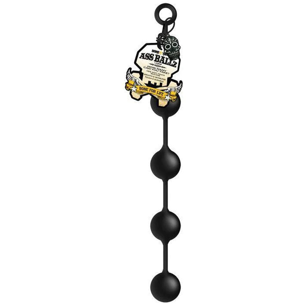 The Boneyard Ass Ballz L - Black 46 cm features soft-touch black silicone anal beads with four decreasingly sized round beads and a circular loop at the top for easy handling. A product tag shows branding and decorative elements partially visible.