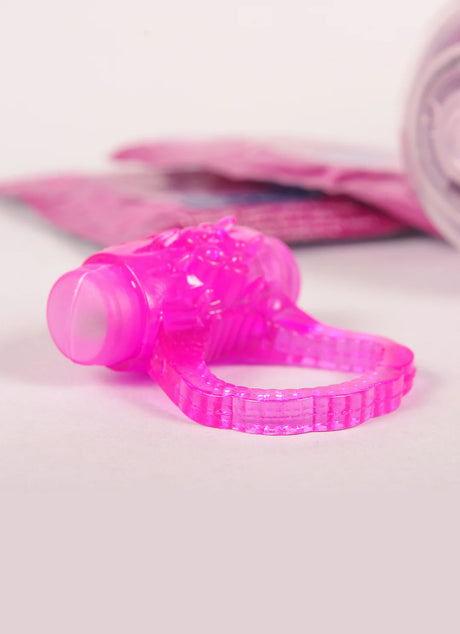 A close-up image of a pink, textured silicone ring with a small vibrating motor on top. It is designed for adult use. The background is softly blurred, with hints of pink packaging visible on the left side.