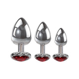 The Adam & Eve Three Hearts Gem Anal Plug Set includes three metallic butt plugs in varying sizes, each with a heart-shaped red gemstone base. Displayed on a white background, the jeweled bases and reflective metal surfaces highlight their glossy finish.