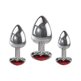 The Adam & Eve Three Hearts Gem Anal Plug Set includes three metallic plugs with reflective silver surfaces and heart-shaped bases adorned with red gems. Displayed against a plain white background, the sizes increase gradually from left to right.