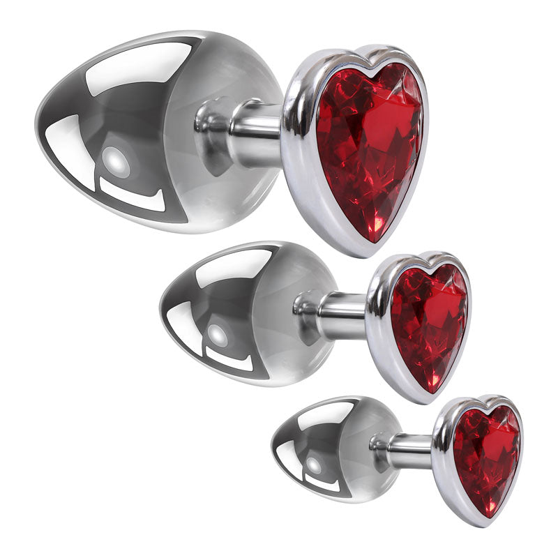 The Adam & Eve Three Hearts Gem Anal Plug Set includes three metallic butt plugs with heart-shaped red gemstone bases, arranged by size. Each plug shines brilliantly, reflecting light on a white background, while the faceted gemstones add sparkle.