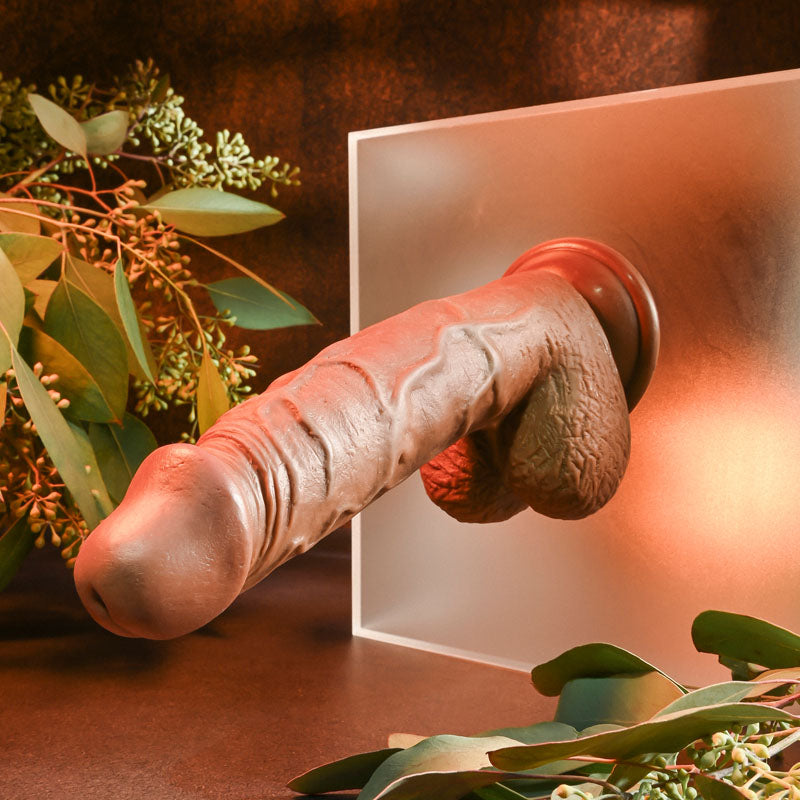 The Evolved Realistic Dildo 8 Dark features lifelike texture and pronounced veins, secured by a powerful suction cup on frosted glass. Green foliage on a dark brown background and soft reddish lighting create a warm ambiance.