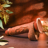 The Evolved REALISTIC DILDO 8 DARK - Brown 21 cm Dong, resembling a lifelike chocolate sculpture, is elegantly displayed on a dark surface. Ambient lighting with green leafy branches and a lit candle set the scene, as this detailed piece rests gracefully in its warm, reddish-brown environment.