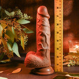 The Evolved REALISTIC DILDO 8 DARK - Brown stands upright on a dark surface next to a measuring tape, surrounded by green leaves and a lit candle, suggesting a natural or rustic theme.