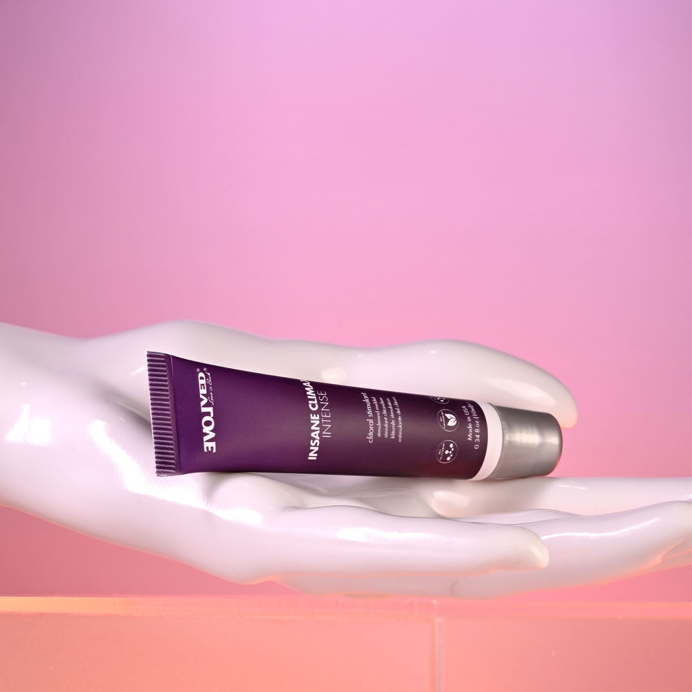 A shiny white mannequin hand holds a dark purple tube labeled Evolved INSANE CLIMAX - INTENSE against a pink gradient. Designed for intimate pleasure, the clitoral stimulation gel has a silver cap and feature icons, with soft illumination adding a reflective glow to the scene.