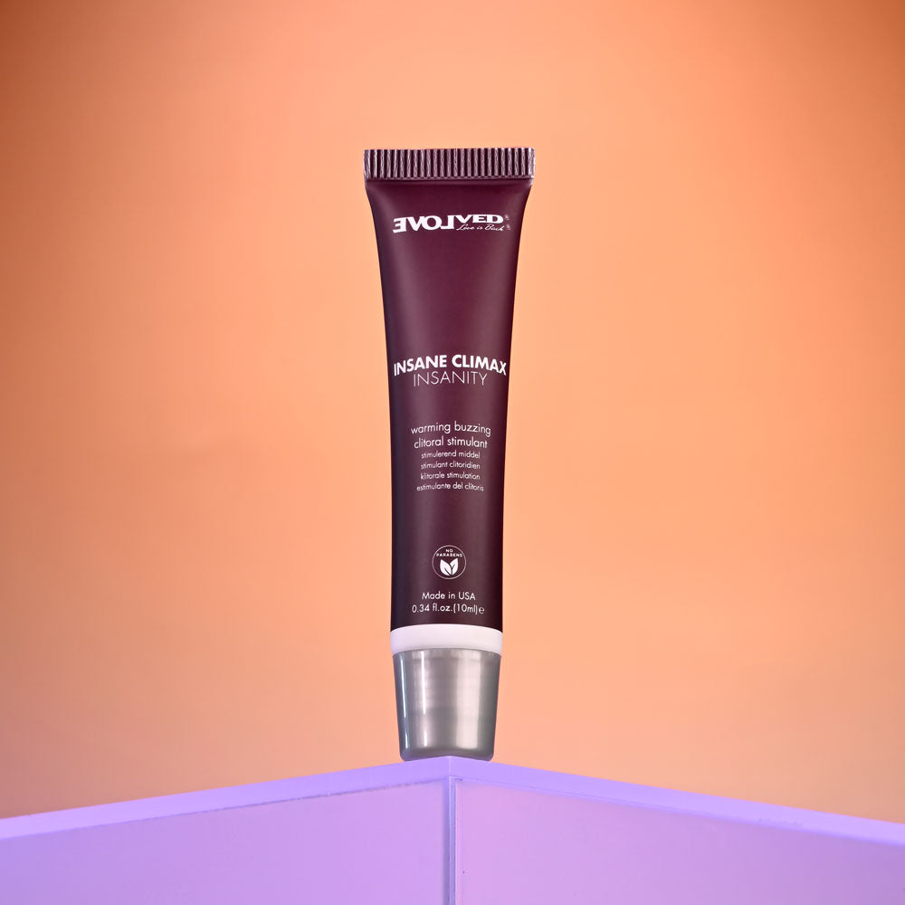 A maroon tube of Evolved INSANE CLIMAX - INSANITY stands upright on a lilac platform against an orange gradient backdrop. Featuring white text and a metallic cap, it offers a warming sensation for enhanced clitoral stimulation, containing 10 ml of gel.