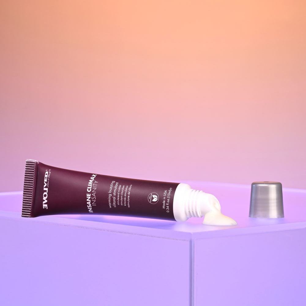 A maroon tube of Evolved INSANE CLIMAX Warming Buzzing Gel rests on a purple surface. The cap, silver and transparent, lies beside it. Squeezing the tube releases some gel, offering a tingle of warmth. The background features a warm orange-pink gradient.