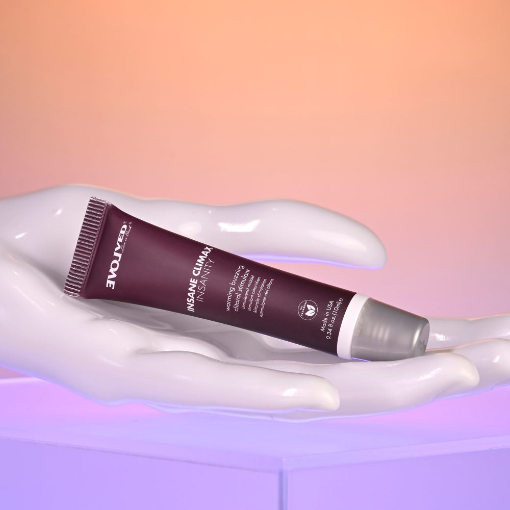 A dark purple tube labeled EVOLVE and containing the Evolved INSANE CLIMAX Clitoral Stimulation Gel rests on a glossy white hand sculpture. A gradient background and pink-lit base hint at the warming sensation of the product.