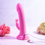 The Evolved SOMEBUNNY TO LOVE Pink Rabbit Vibrator stands upright on a marble surface, connected to its USB charging cable. With pink roses and white flowers softly blurred in the background, it creates a romantic scene ideal for its waterproof design.