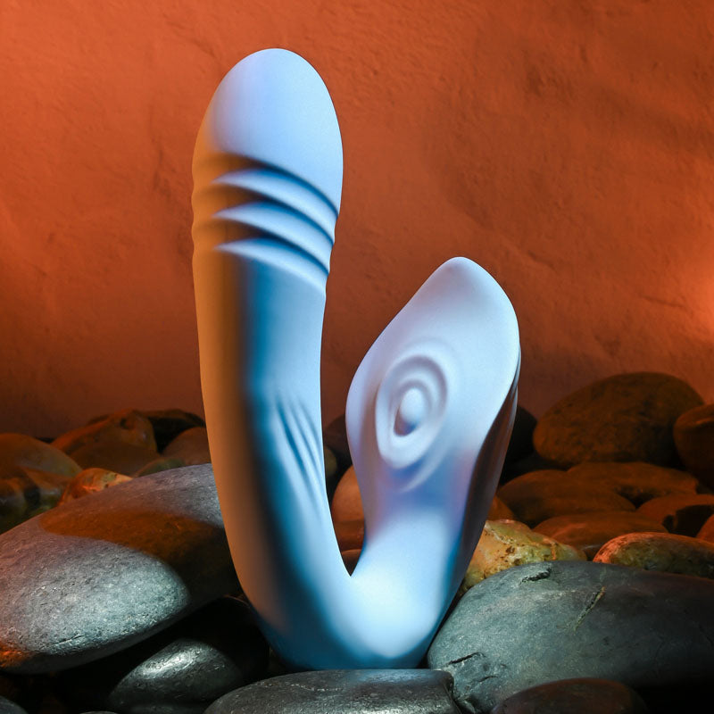 The Evolved TAP & THRUST, a light blue USB rechargeable dual vibrator, features a ribbed texture on one end. It stands elegantly on smooth rocks against an orange-lit backdrop, showcasing its sleek design and textured curves in a warm ambiance.