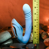 The Evolved TAP & THRUST - Blue USB Rechargeable Dual Vibrator, made of light blue silicone with textured ridges and three buttons at its base, stands on smooth stones beside a yellow measuring tape showing 7 inches. A soft orange glow in the background enhances its elegant allure.