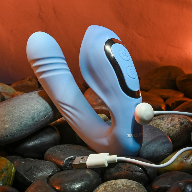 The Evolved TAP & THRUST, a blue USB rechargeable dual vibrator with two prongs and front buttons, rests on smooth gray stones. A USB charging cable is attached, and the background glows warmly in orange.