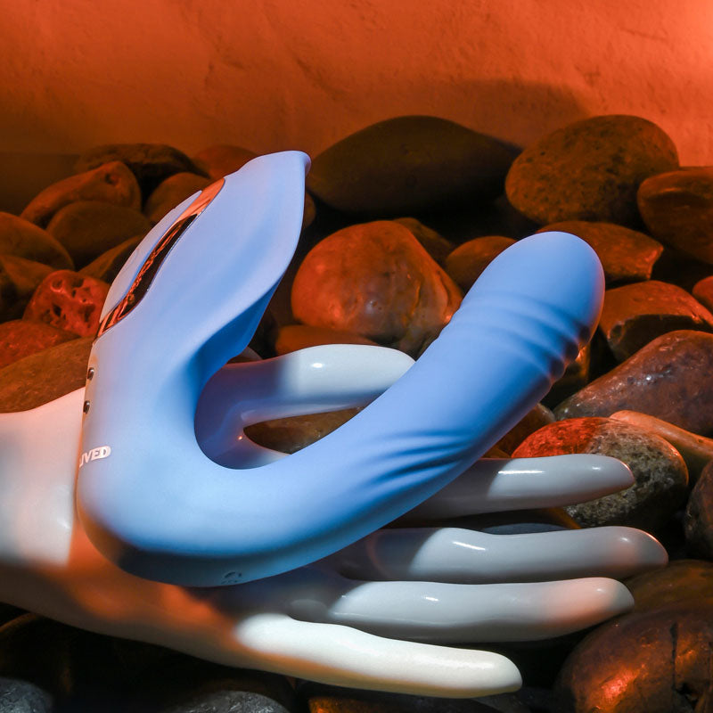 An Evolved TAP & THRUST – Blue USB Rechargeable Dual Vibrator, featuring buttons on the side, rests on a white mannequin hand. Its elegantly placed on smooth stones with a warm reddish glow background that highlights their texture.