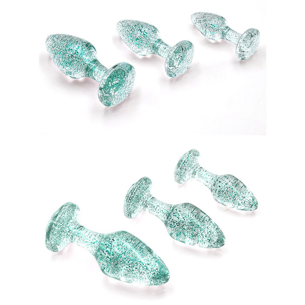 The Adam & Eve Glitter Gem 3-Piece Butt Plug Set features three transparent glass plugs with shimmering turquoise flecks. Displayed on a white background, they vary in size: small, medium, and large, with tapered tips and gem-shaped bases for optimal anal training.