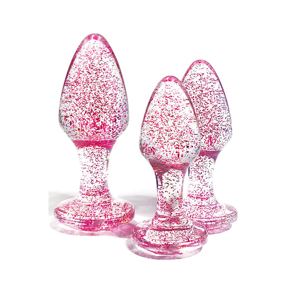 The Adam & Eve Glitter Gem 3-Piece Butt Plug Set features translucent pink glass plugs with tapered, bulbous shapes and round bases. Speckled with metallic glitter for sparkle, these decorative pieces vary in size and stand upright on a white background.