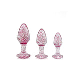 Three teardrop-shaped plugs from the Adam & Eve Glitter Gem 3-Piece Butt Plug Set, made of shimmering material with pink speckles, are artistically arranged from smallest to largest; their flared bases have a decorative gem shape, all set against a white background.