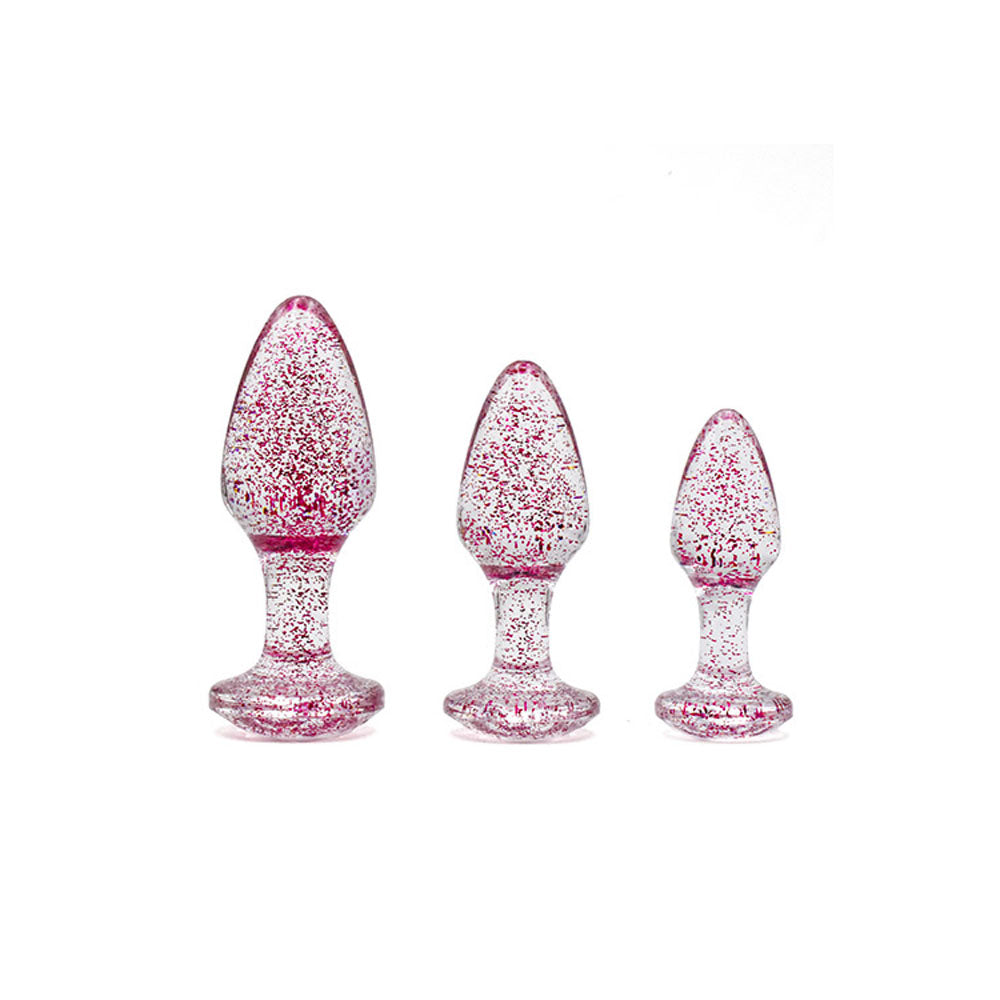 The Adam & Eve Glitter Gem 3-Piece Butt Plug Set in pink features three transparent glitter plugs with tapered tips and flared bases, arranged from large to small against a plain white background, ideal for anal training.