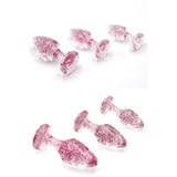 The Adam & Eve Glitter Gem 3-Piece Butt Plug Set features pink, glittery, translucent plugs in three sizes with tapered shapes and gem-shaped bases. Displayed on a white background, the overhead view highlights their sparkle while the side view showcases their elegant form for anal training.