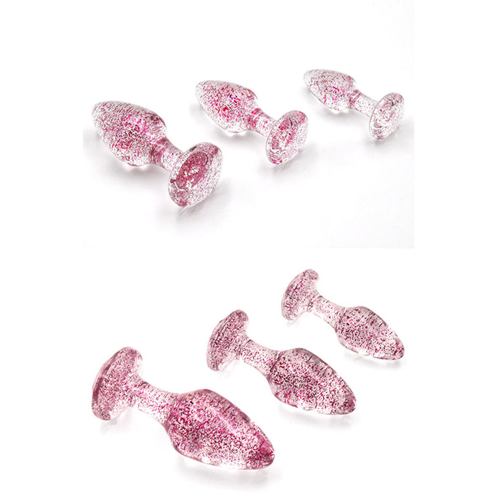 The Adam & Eve Glitter Gem 3-Piece Butt Plug Set includes small, medium, and large pink-glitter plugs displayed in front and side views, with textured glass and pink specks enhancing their decorative charm.