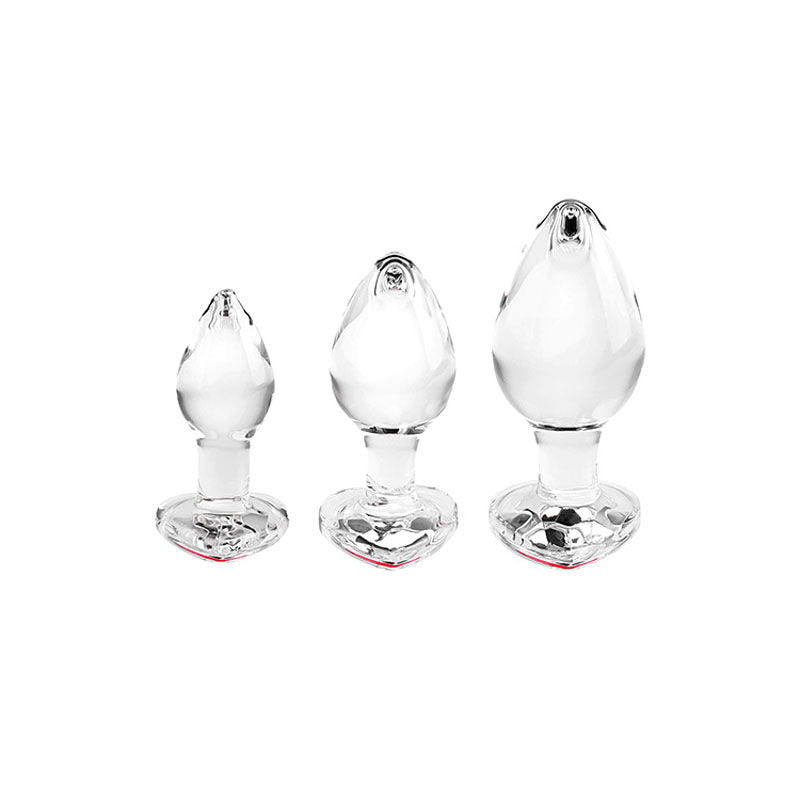 The Adam & Eve RED HEART GEM GLASS PLUG SET features three clear glass butt plugs, each with a red jewel heart base. Elegantly lined up by size on a white background, this set is perfect for anal training enthusiasts.