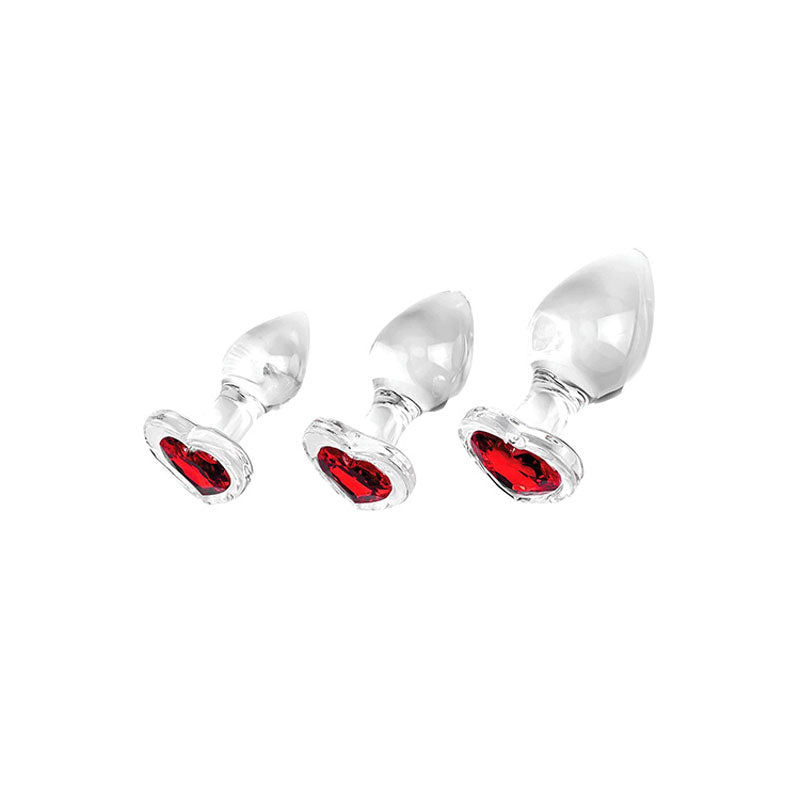 The Adam & Eve RED HEART GEM GLASS PLUG SET features three clear glass butt plugs with heart-shaped red gem bases, offering small to large sizes for anal training. Their tapered design and transparent finish beautifully contrast with the vibrant red gems.