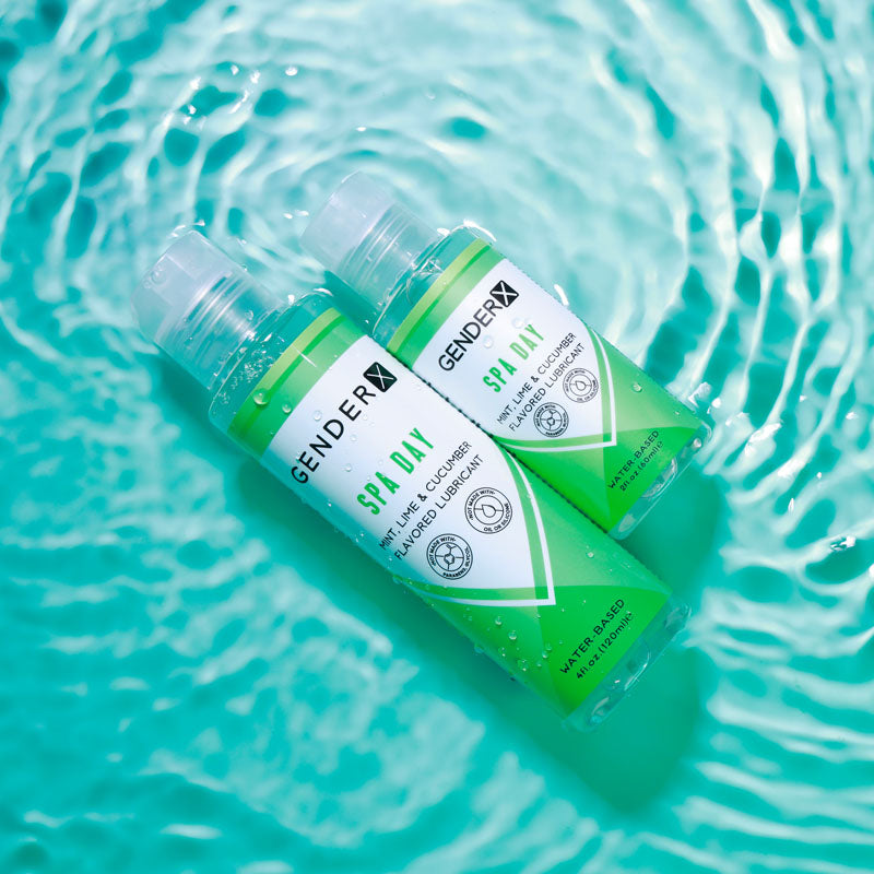 Two green bottles of Gender X SPA DAY Flavoured Lube, a mint, lime, and cucumber water-based lubricant, float on rippling blue water. The bottles feature white labels with geometric designs and transparent caps. Sunlight highlights both the textures of the water and bottles, creating a relaxing atmosphere.