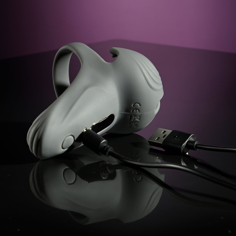 The Gender X UNDERCARRIAGE is a gray, textured silicone vibrating ring resting on a glossy black surface with several cutouts. Two black cables, one USB, connect to it. Its waterproof design contrasts against a dark to light purple gradient.