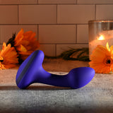 A Gender X ANYBODYS PLUG in blue silicone rests on a gray stone countertop, with orange flowers and candlelight creating a warm ambiance. This waterproof vibrating butt plug complements the cozy setting framed by white subway tiles, adding an intimate touch.