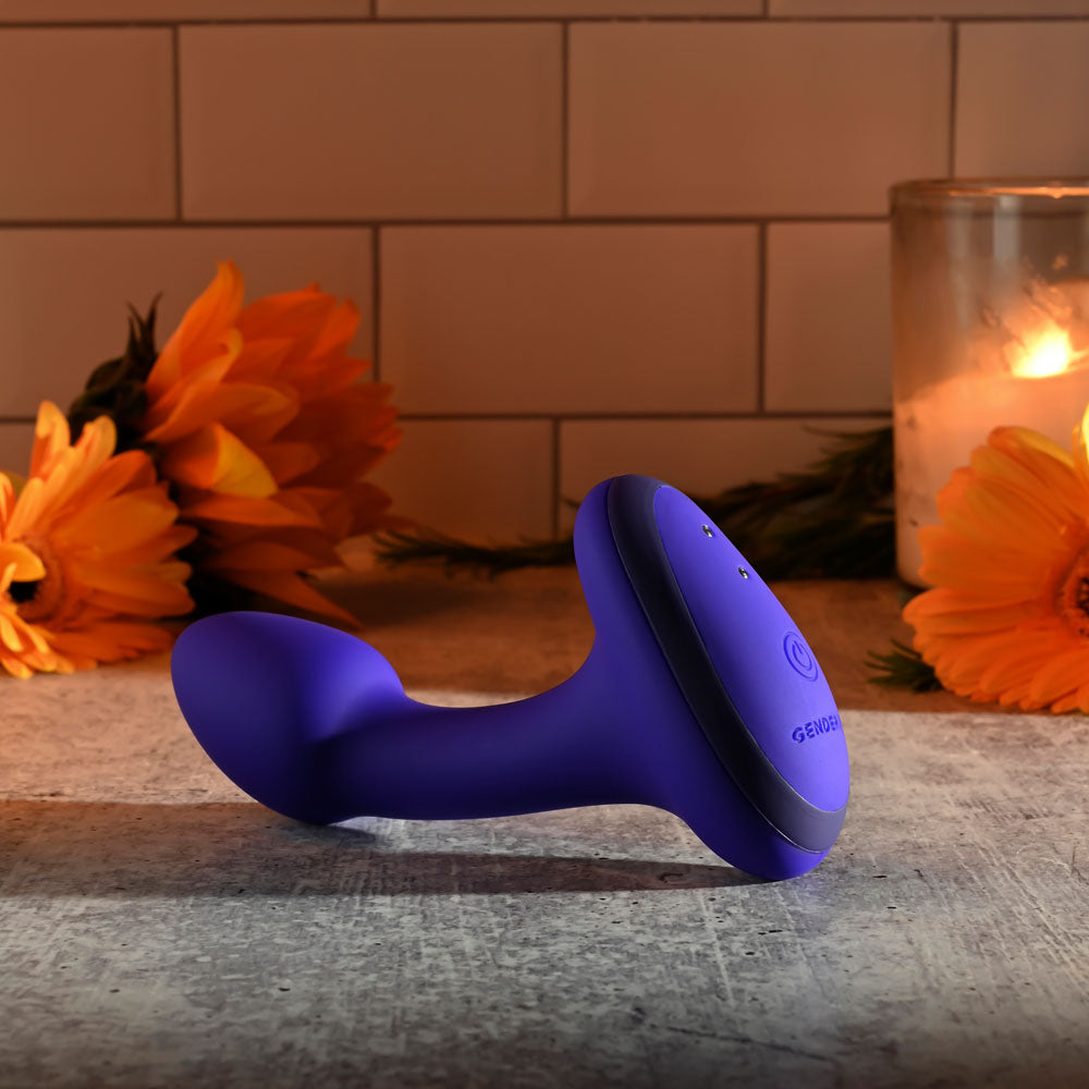 A blue, curved Gender X ANYBODYS PLUG, crafted from silicone, rests on a stone surface lit by candlelight. Two lit candles and three orange flowers enhance the serene ambiance against a warm white tiled wall in this tranquil setting.