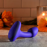 A blue, L-shaped Gender X ANYBODYS Plug with a power button rests on a textured surface. In the background, a lit candle and orange flowers enhance the inviting atmosphere. With multiple vibrating speeds, it brings excitement to any setting against the tiled wall backdrop.