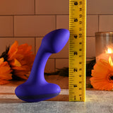 A blue Gender X ANYBODYS PLUG vibrating butt plug, approx. 11.4 cm long, stands upright beside a yellow ruler for scale on a textured surface with orange flowers and a lit candle in the background. The white-tiled wall enhances the cozy ambiance.