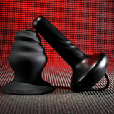 A black silicone adult toy is displayed against a textured red-hued backdrop. It features the Gender X ECHO - Black 15.3 cm USB Rechargeable Butt Plug with a smooth, cylindrical design and wide base. The background highlights its sleek, matte black finish.