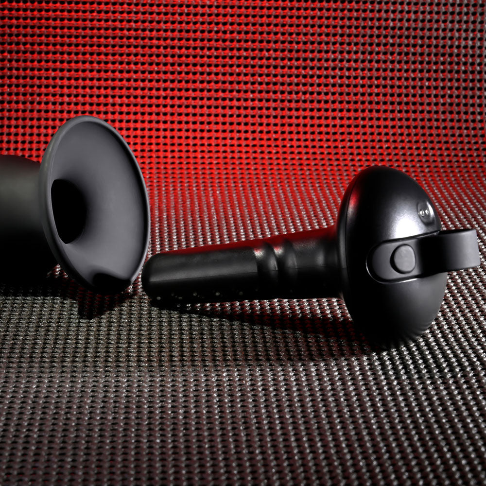 A black Gender X ECHO USB rechargeable butt plug, featuring a 15.3 cm silicone design for added grip, rests elegantly on a textured surface with a red grid pattern. Positioned at an angle, it casts intriguing shadows, its smooth surface accentuating the modern design.