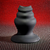 The Gender X ECHO - Black 15.3 cm USB Rechargeable Butt Plug features a sculptural, tiered design with smooth, wavy curves. Standing gracefully on a textured reddish surface and set against a red mesh backdrop, the lighting highlights its matte finish and elegant form.