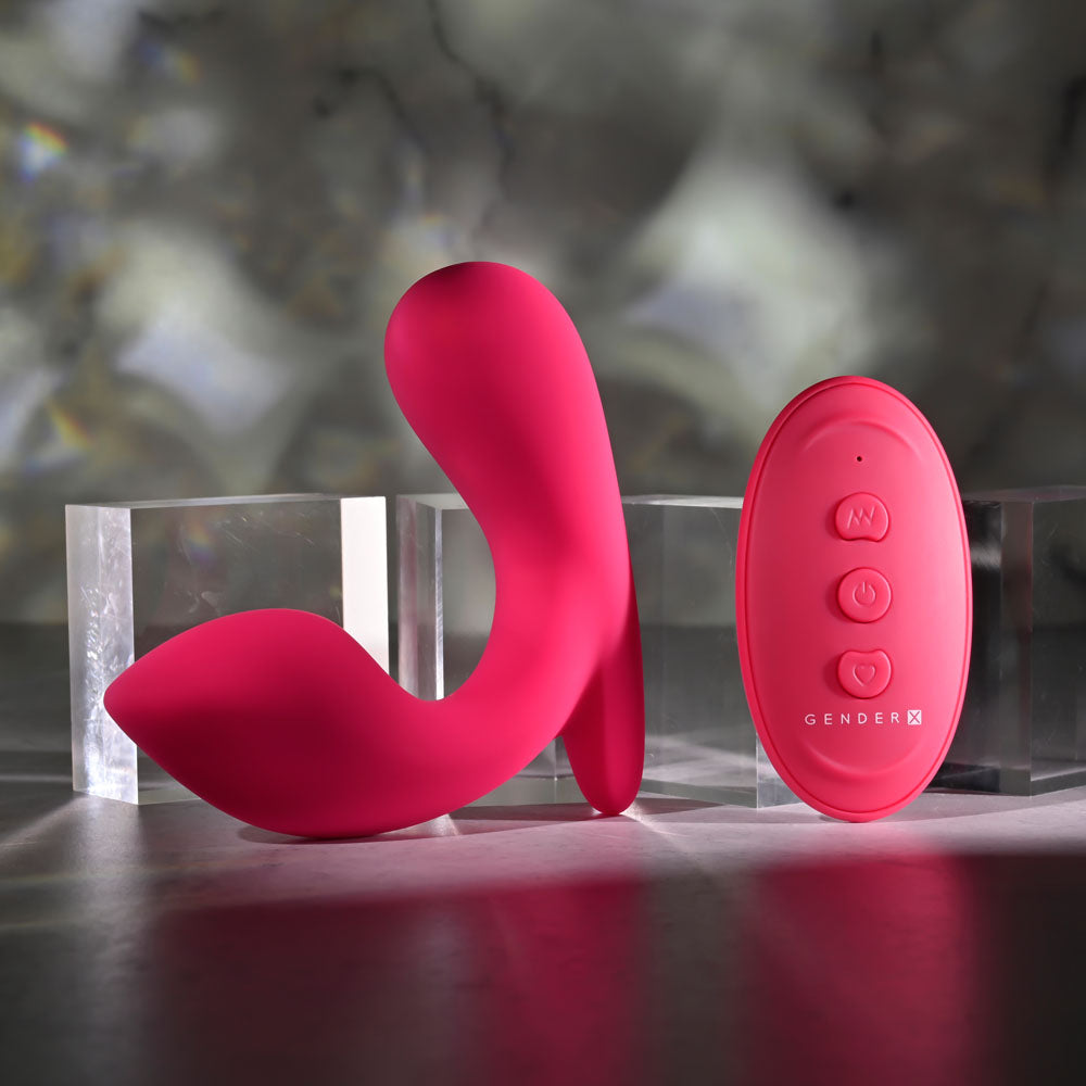 The Gender X ALL ABOUT THE BASS, a pink wearable vibrator with an ergonomically designed silicone body and a wireless remote control, stands upright on a reflective surface next to two clear acrylic blocks. The backdrop features blurred gray and white tones for a soft, abstract effect.