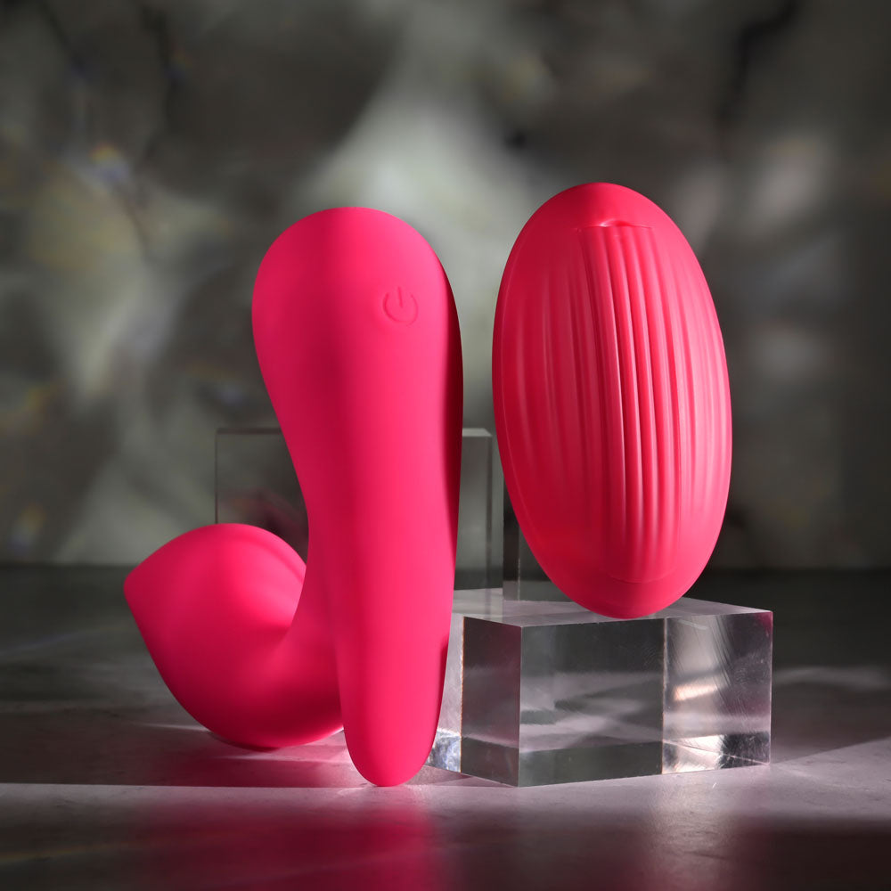 Two pink, modern Gender X ALL ABOUT THE BASS vibrating toys are displayed on clear acrylic stands. The left, crafted from silicone and curved with a power symbol, contrasts with the oval one featuring vertical ridges. Soft lighting highlights their bright color and remote control features against a dark background.