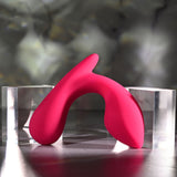 A sleek, pink Gender X ALL ABOUT THE BASS vibrator, resembling an abstract sculpture, arcs between two transparent blocks. The softly blurred gray background and subtle lighting highlight its smooth texture and vibrant color.