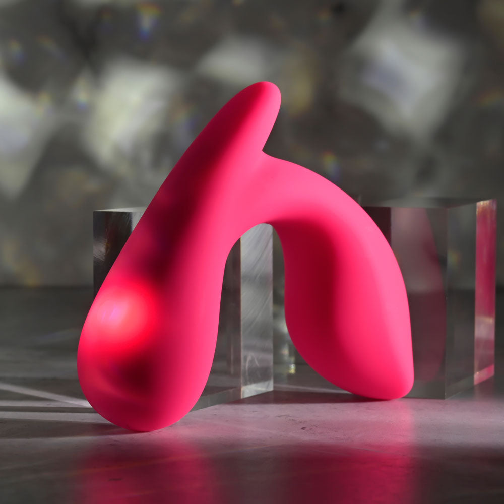 The Gender X ALL ABOUT THE BASS, a pink, abstract U-shaped wearable vibrator with smooth silicone surfaces, is placed on a table beside transparent rectangular blocks. A light source casts soft shadows on it against a neutral, blurred background.