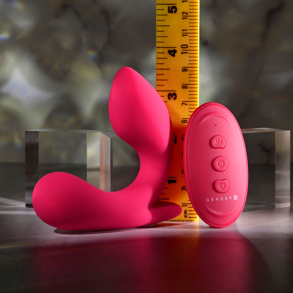 The Gender X ALL ABOUT THE BASS is a pink, dual-tipped silicone USB rechargeable wearable vibrator with a wireless remote. Its shown next to a yellow measuring tape for size comparison and surrounded by reflective glass cubes on a gray surface.