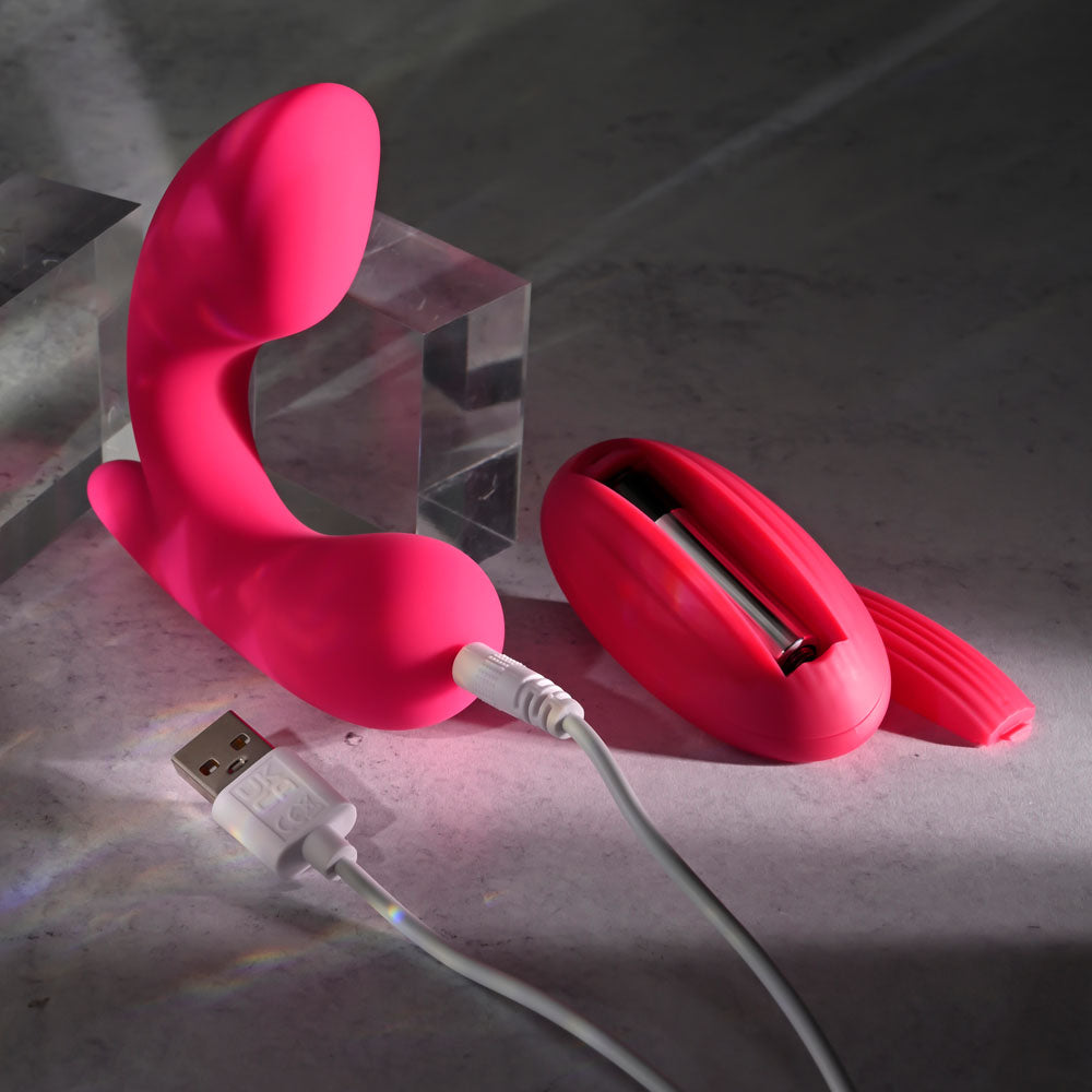 The Gender X ALL ABOUT THE BASS is a pink, curved silicone G-spot vibrator with a USB charging cable. It rests on a gray surface alongside a matching remote control, both highlighted by soft lighting. Transparent acrylic blocks in the background add a modern touch to the ensemble.