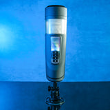 The Gender X MESSAGE IN A BOTTLE, a sleek cylindrical device with a black and metallic finish, rests on a stand. An illuminated button panel in the center activates turbo mode. The background features a blue gradient, with the surface reflecting its base.